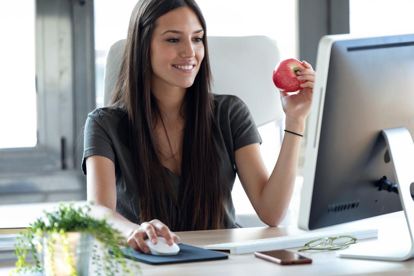 Junge hübsche Frau die einen Apfel ist und mit einem Lächeln ihre Bewerbung an einem Apple iMac an ZENNER Ventilatoren sendet